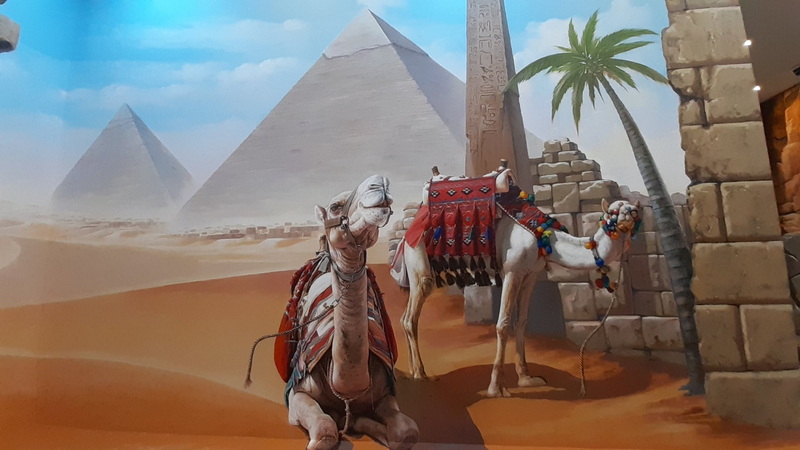 Camel statues with colorful accessories against a desert scene with pyramids and a palm tree at Chiang Mai Art in Paradise.
