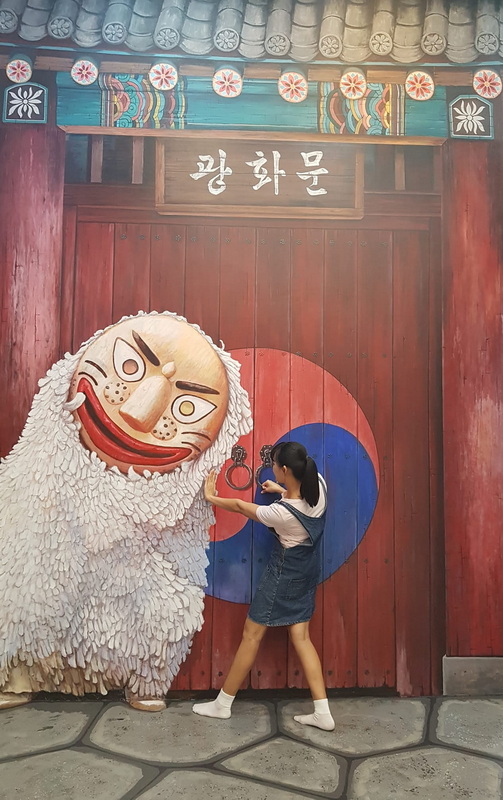 Girl holding a key-shaped object in front of a large mask sculpture on a traditional door at Art in Paradise Chiang Mai.