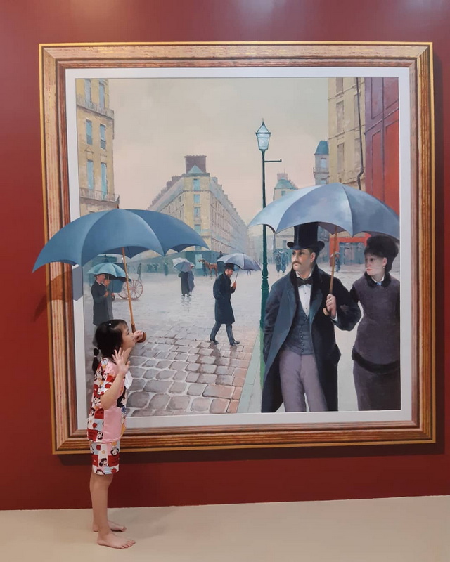 Child looking at painted street scene with umbrellas and historical-dressed figures at Art in Paradise Chiang Mai.