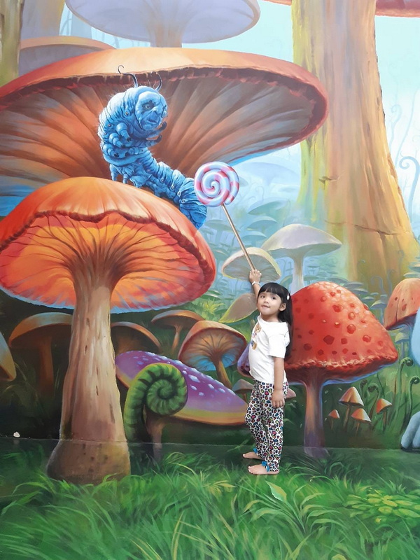 Fantasy Mushroom Mural Scene at Art in Paradise Chiang Mai Child posing in front of a vibrant fantasy mural with giant mushrooms and a blue caterpillar at Art in Paradise Chiang Mai.