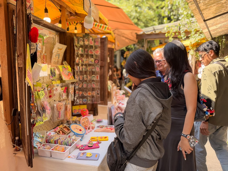Outdoor Market Stall Shopping at Baan Kang Wat People shopping at an outdoor market stall with colorful crafts and accessories on display at Baan Kang Wat.