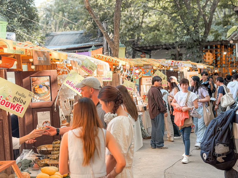Crowd shopping at a vibrant outdoor market with colorful signs and trees at Baan Kang Wat.
