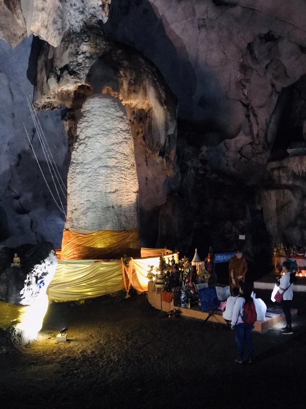 Large stalagmite inside a cave decorated with cloth and small statues, illuminated by lights at Mueang On Cave.