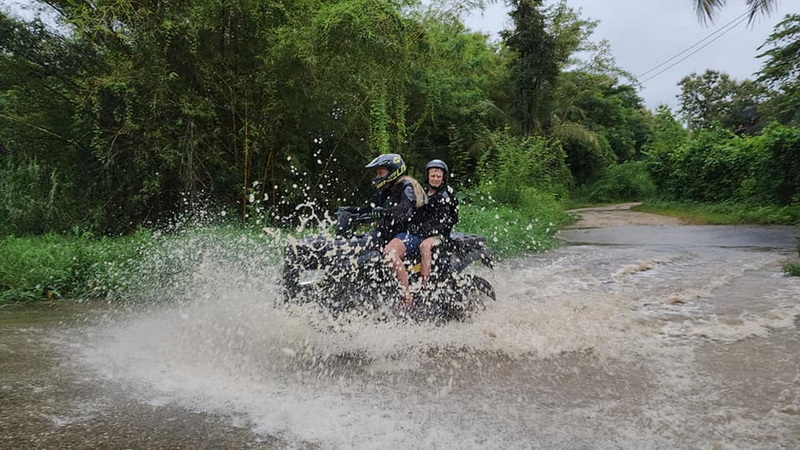 Two people riding ATV through shallow water on a trail surrounded by greenery at ATV tour Chiang Mai.