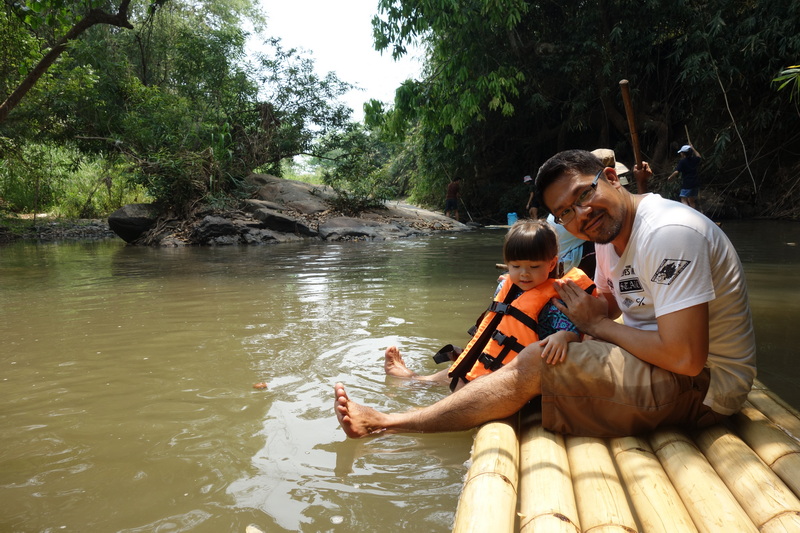 Man and girl sitting on a bamboo raft in a muddy river with lush greenery around at Bamboo Rafting in Mae Wang.