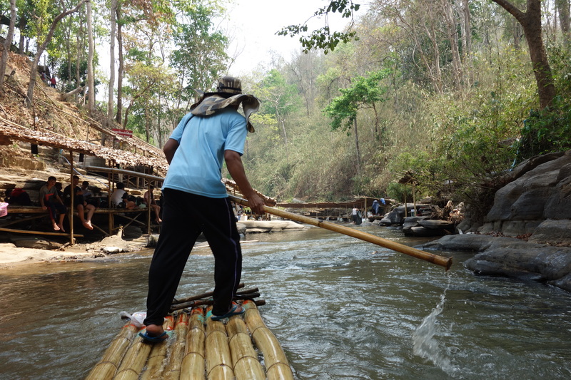 Person navigating a bamboo raft on a river with trees and rocks around at Bamboo Rafting in Mae Wang.