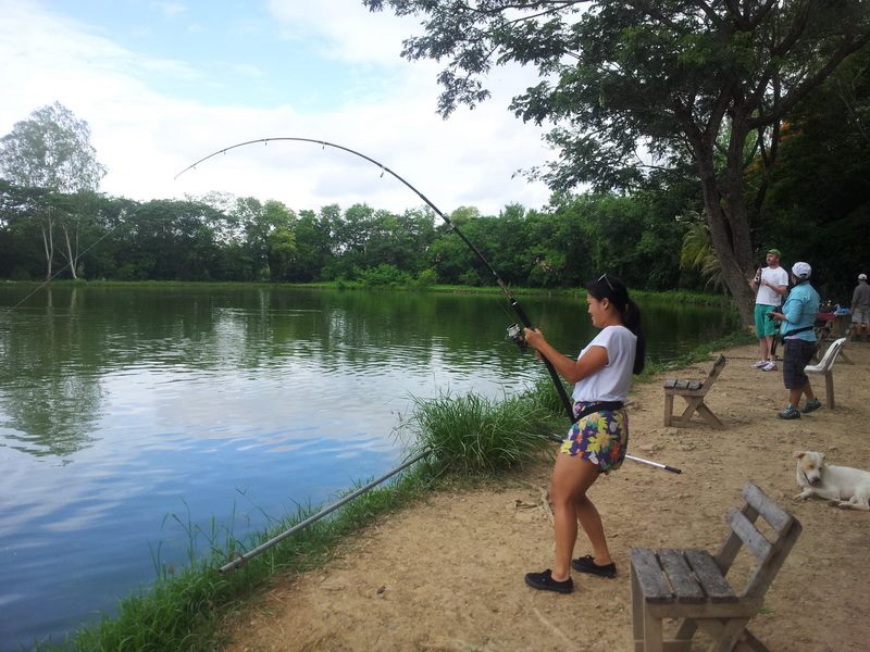 Woman fishing by the river surrounded by trees and other people at Chiang Mai Fishing Park.