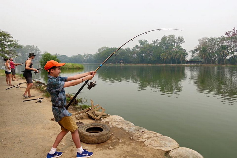 Boy fishing with a rod on the riverbank as others fish nearby at Chiang Mai Fishing.
