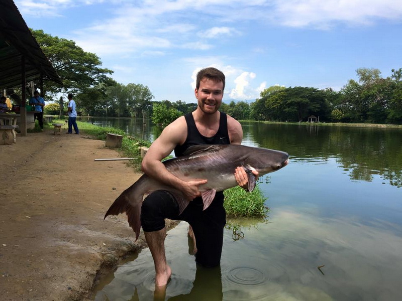 Man holding a large fish by the riverbank with trees and sky in the background at Chiang Mai Fishing.