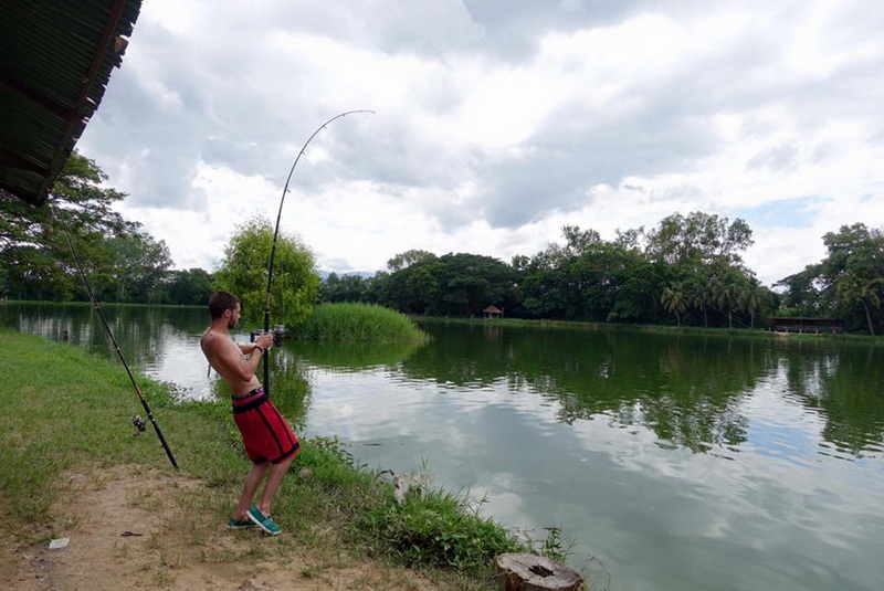 Boy fishing by a tranquil river with trees and cloudy sky in the background at Chiang Mai Fishing.