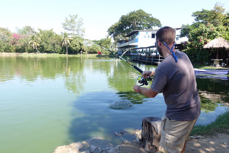 Man fishing by a tranquil lake with trees and buildings behind him at Chiang Mai Fishing Park.