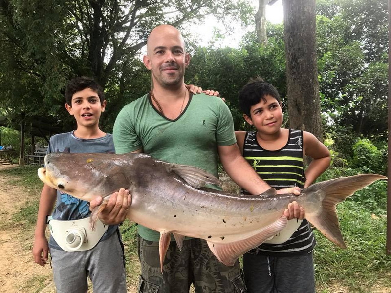 Family with Large Fish outdoors at Chiang Mai Fishing Man and two children with large catfish in outdoor setting surrounded by trees at Chiang Mai Fishing.