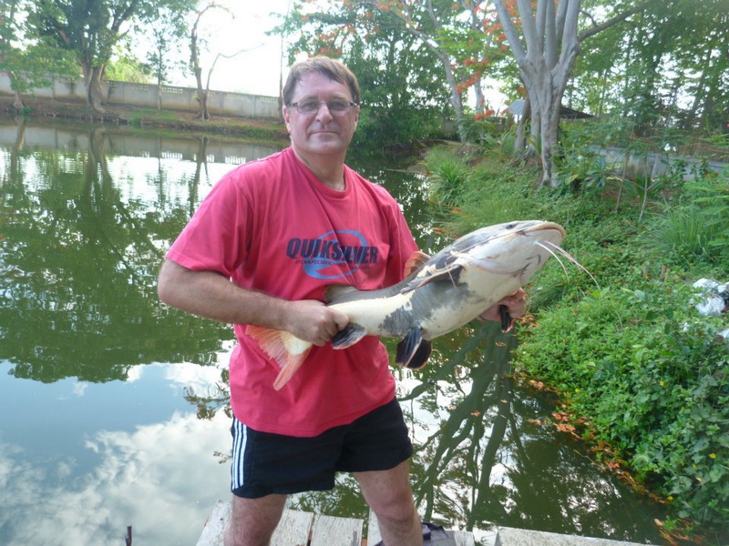 Fishing Success by the Pond at Chiang Mai Fishing Park Man holding a large catfish near a pond surrounded by trees and greenery at Chiang Mai Fishing Park.