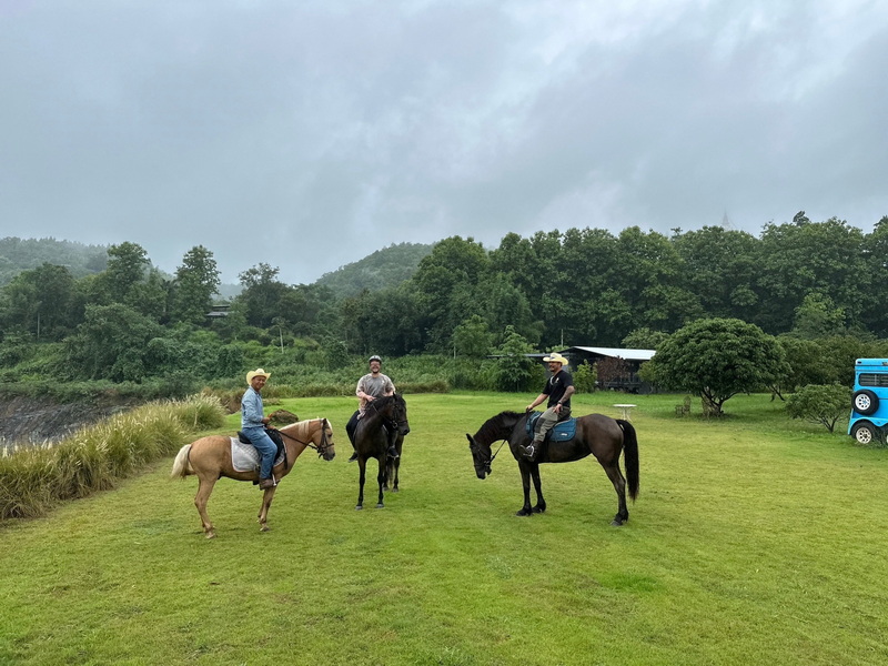 Three riders with horses on a grassy field surrounded by trees and hills at Horse Riding Chiang Mai.