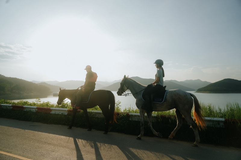 Two riders on horses walking by a lakeside with mountains and sky in background at Horse Riding Chiang Mai.