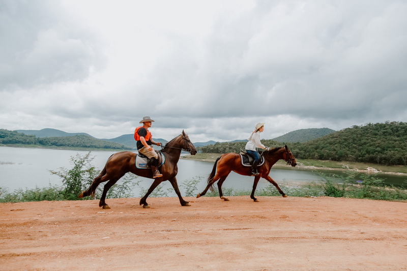 Two riders on horses traveling along a dirt trail by a lake with hills and a cloudy sky at Horse Riding Chiang Mai.