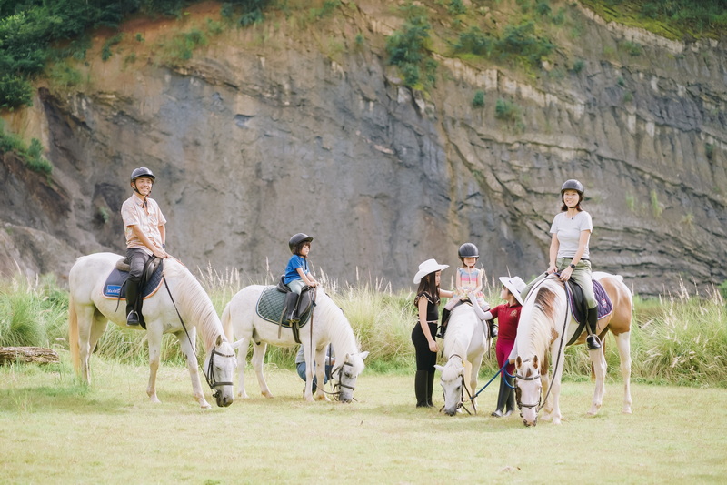 Group of children and adults riding horses on grassy field with cliffs behind at Horse Riding Chiang Mai.