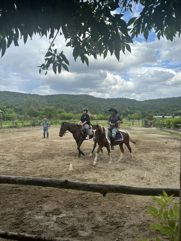 Horse Riding Scene at Horse Riding Chiang Mai Two riders on horses in an outdoor arena with mountains and trees in the background at Horse Riding Chiang Mai.