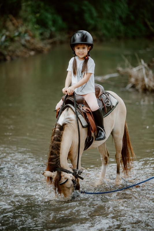 Young girl riding a horse through a river with green trees around at Horse Riding Chiang Mai.