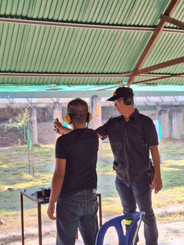 Two men at a shooting range under a metal roof, one helping the other with a gun at Chiang Mai Shooting Club.