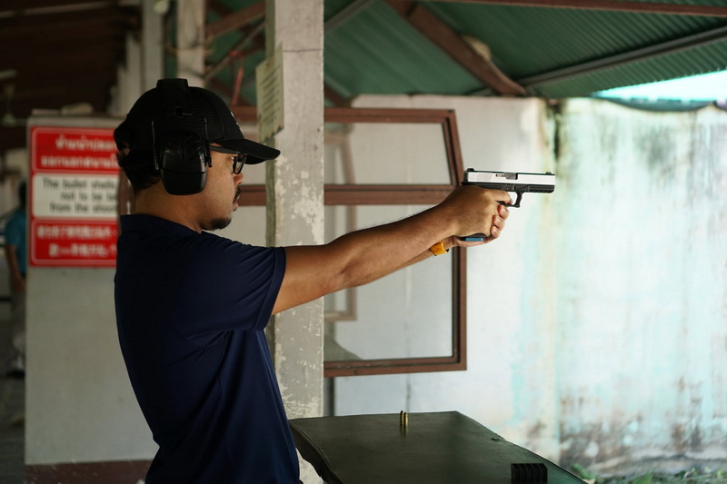 A man in headphones aiming a handgun at a shooting range with walls and windows behind him at Chiang Mai Shooting Club.