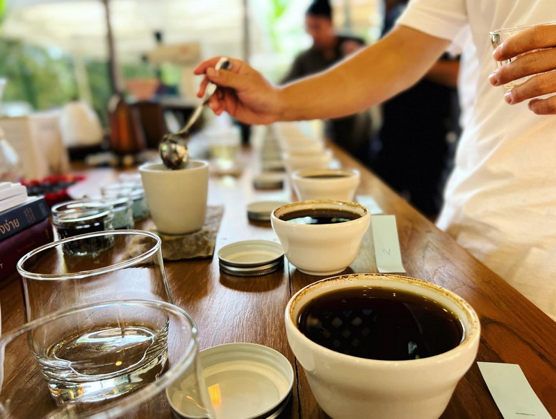 Close-up of black coffee cups and glasses on a wooden table with people in the background at Chiang Mai Coffee Farm Tour.