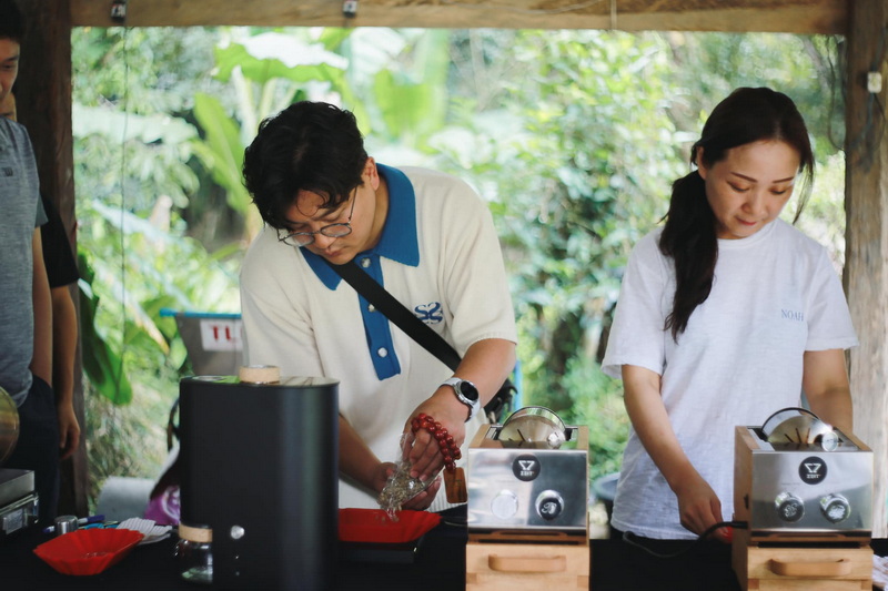 Outdoor Coffee Preparation Scene at Chiang Mai Coffee Farm Tour Two individuals preparing coffee outdoors with green plants behind them at Chiang Mai Coffee Farm Tour.