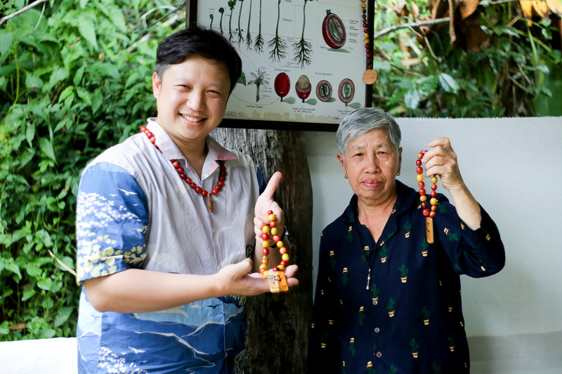 Two women holding colorful beaded necklaces outdoors with greenery and plant root diagram behind them at Coffee Farm Tour Chiang Mai.