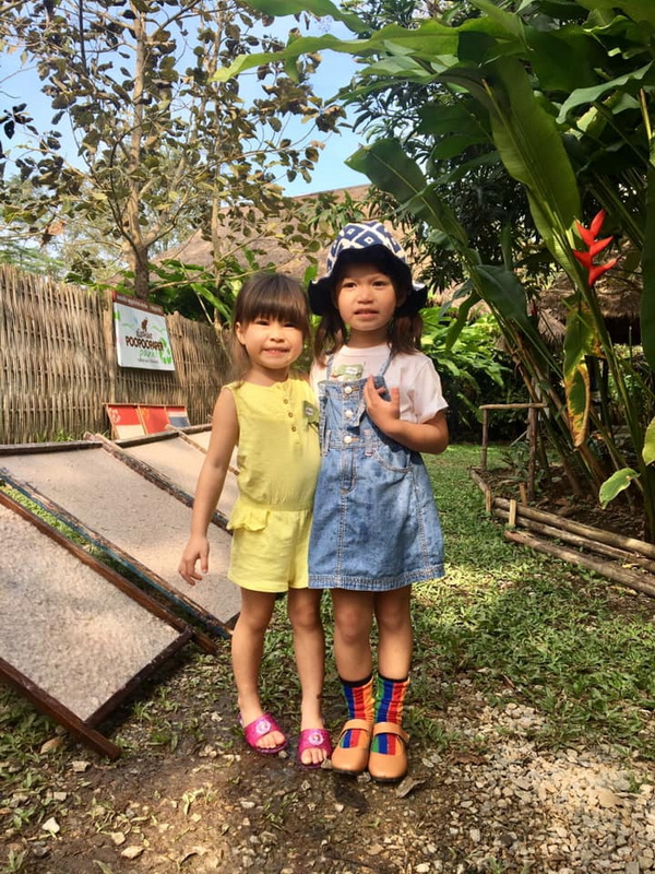 Two girls in colorful outfits standing on a dirt path with plants and a fence in the background at Elephant Poopoo Paper Park.