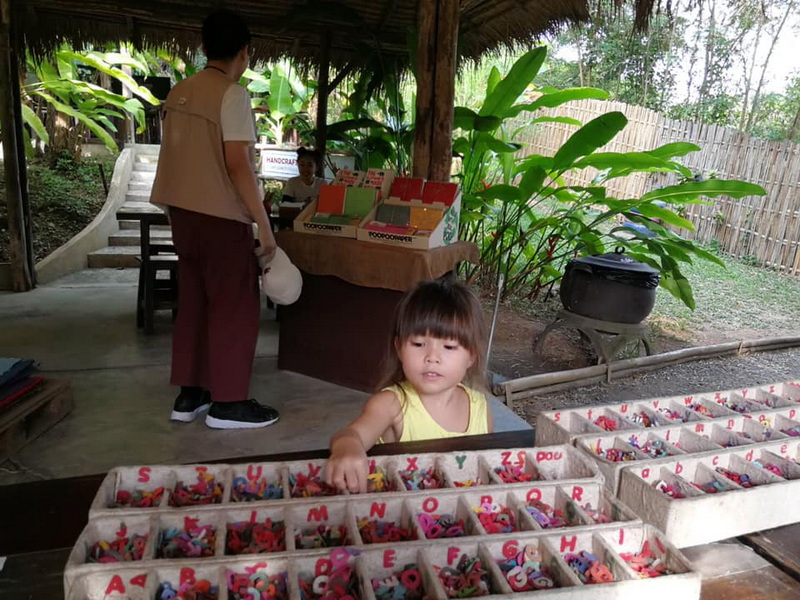 Girl with Beads in a Shelter at Elephant Poopoo Paper Park Young girl reaching for colorful beads in a wooden display inside a sheltered area with plants outside at Elephant Poopoo Paper Park.