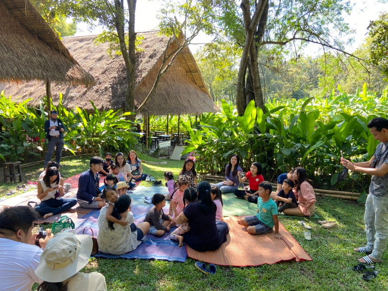 Outdoor Group Gathering at Elephant Poopoopaper Park People gathering on mats under trees amid bamboo huts and lush greenery in an outdoor setting at Elephant Poopoopaper Park.