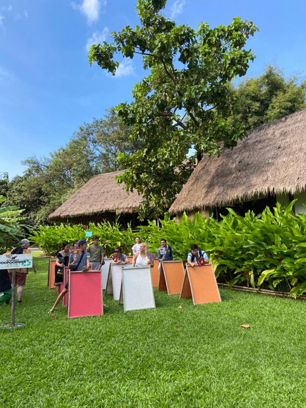 Visitors Outside at Elephant Poopoopaper Park at Elephant Poopoo Paper Park Visitors gather outside next to colorful boards and thatched-roof huts amid greenery at Elephant Poopoo Paper Park.
