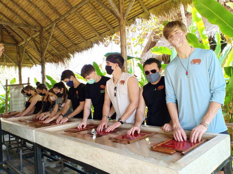 People participating in a craft activity under a thatched roof surrounded by tropical plants at Elephant Poopoopaper Park.
