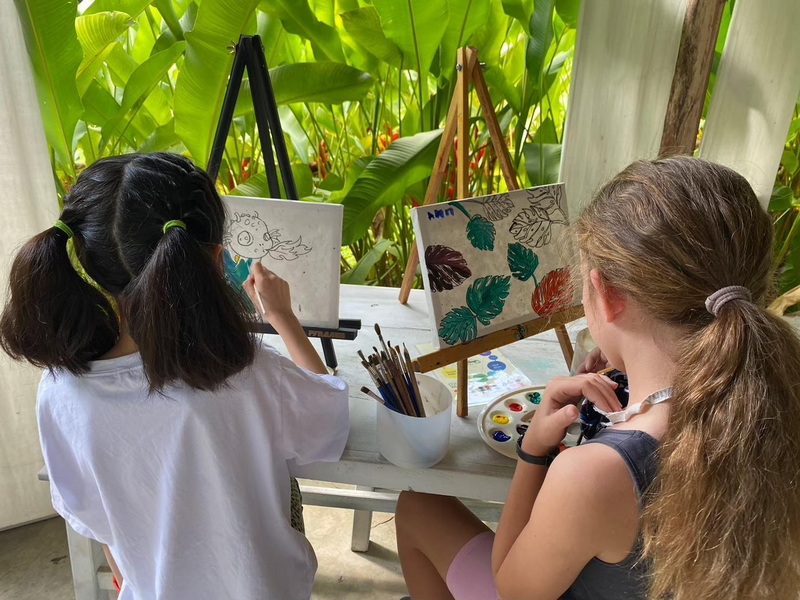 Two girls painting on canvases with green plants in the background at Elephant Poopoo Paper Park.