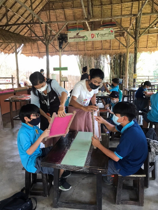 Children and adults making paper crafts under a bamboo pavilion with a sign overhead at Elephant Poopoo Paper Park.