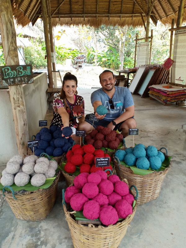 Smiling man and woman surrounded by baskets of colorful felted balls in a rustic shop setting at Elephant Poopoopaper Park.