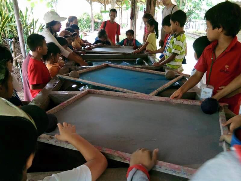 Group of children and adults around sandboxes outdoors with greenery in the background at Elephant Poopoopaper Park.