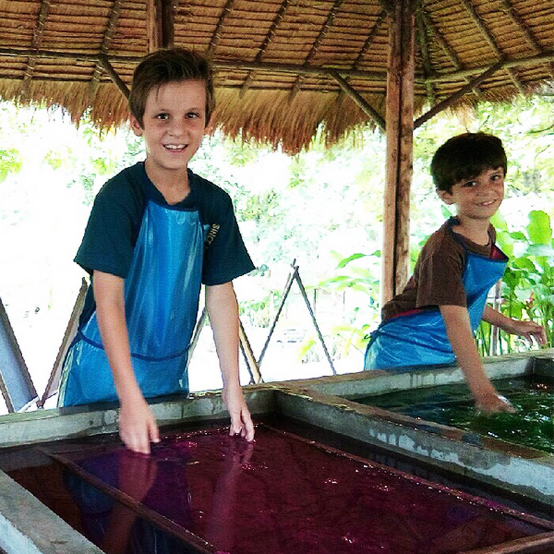 Two children smiling and touching colorful water tanks inside a wooden shelter at Elephant Poopoo Paper Park.