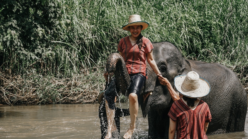 Woman sitting on an elephant in a river with another person nearby holding its trunk at Mae Rim Elephant Home.