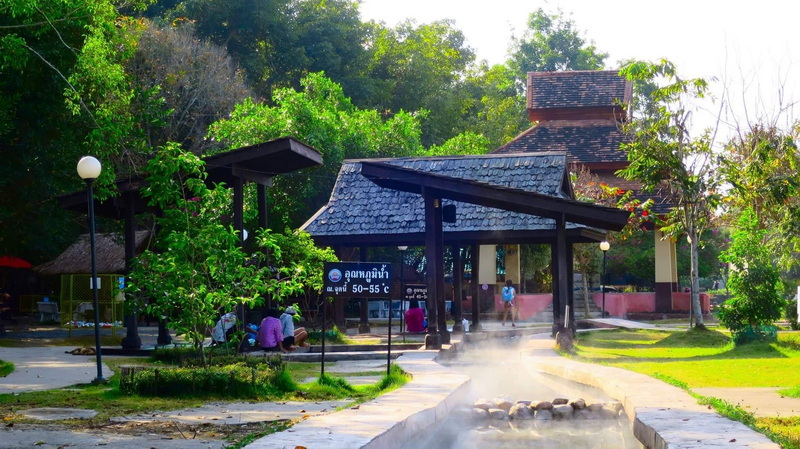 Park with pavilion and steam rising from hot springs surrounded by trees at San Kamphaeng Hot Springs.