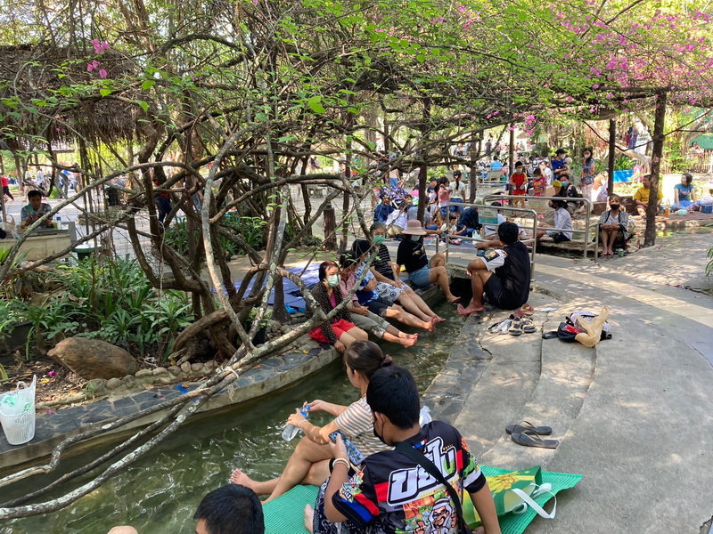 People relaxing by a stream surrounded by trees and pink blossoms in a park at San Kamphaeng Hot Springs.