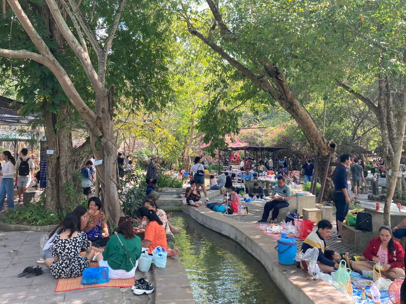 People enjoying a picnic along a waterway under large trees in a park setting at San Kamphaeng Hot Springs.