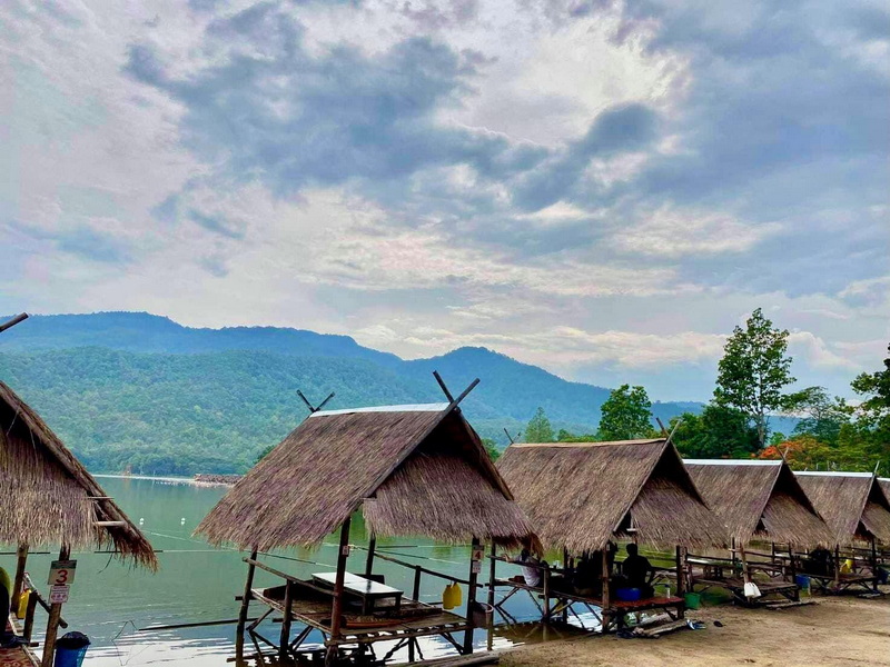 Thatched huts on the lakeshore with mountains and cloudy sky behind at Huay Tung Tao.
