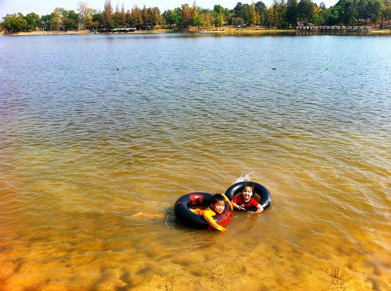 Children playing in a lake with inner tubes, trees and buildings in background at Huay Tung Tao.