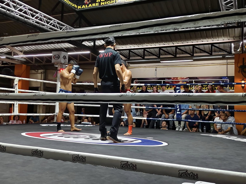 Two Muay Thai fighters in a ring with a referee and audience watching indoors at Kalare Boxing Stadium.