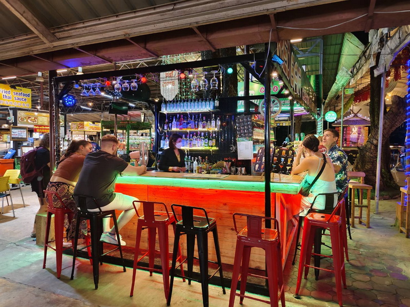 Night Market Bar Scene at Chiang Mai Night Markets People sitting at a lit bar counter inside a lively market area at night at Chiang Mai Night Markets.