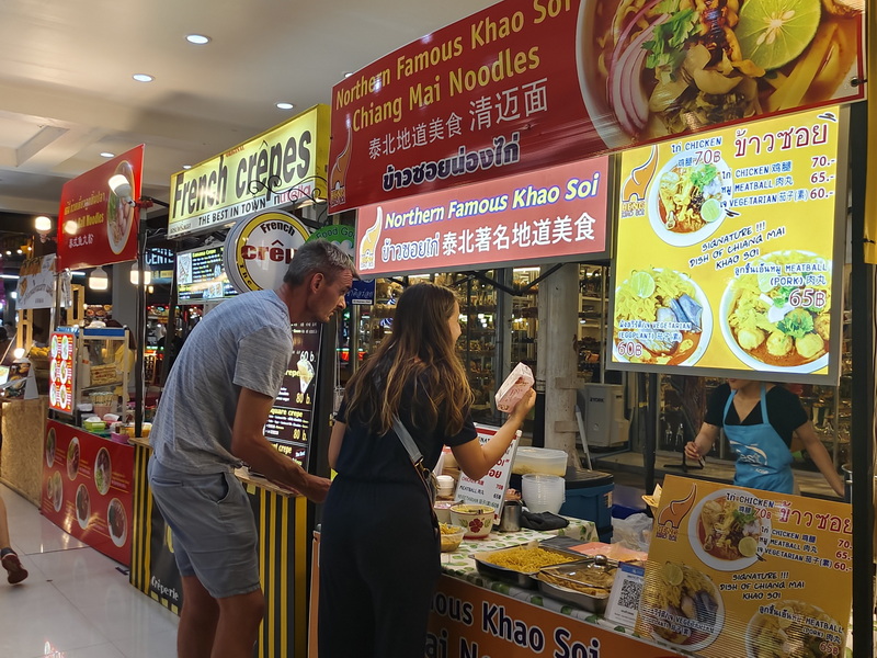 Man and woman buying food at a busy indoor Thai market stall with bright signs at Chiang Mai Night Bazaar.