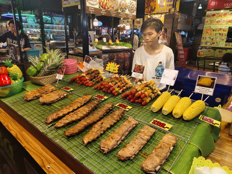 Woman selling grilled meat skewers and corn at a busy market stall at Chiang Mai Night Bazaar.