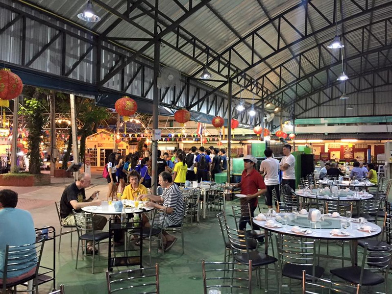 Indoor dining area with tables, chairs, and people, open ceiling with hanging lanterns and outdoor trees at Chiang Mai Night Bazaar.