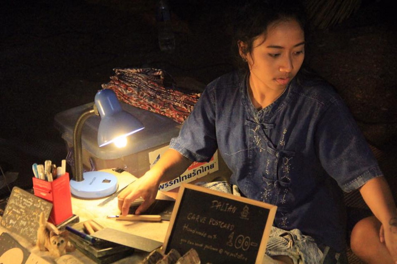 Woman arranging items at a table with a small lamp in a dark environment at Saturday Walking Street Chiang Mai.